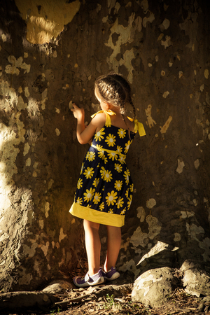 little girl in cute dress stand by huge tree play with bark back shot summer day in parkの写真素材