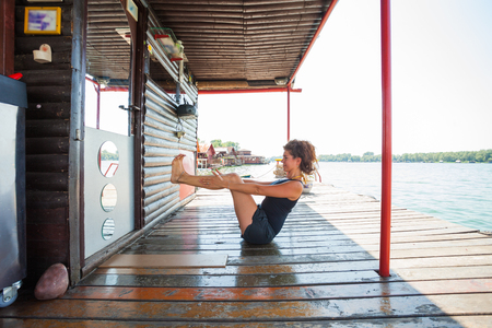 young woman doing exercise for abdominal muscles on wooden river raft by river summer day の写真素材