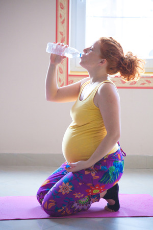red head pregnant young woman on yoga class indoor drinking water from bottleの写真素材