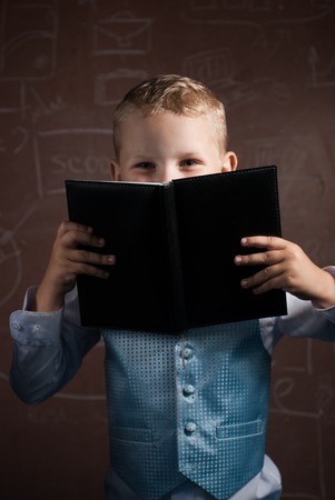 Little schoolboy with blond hair in a nice suit, an emotional student with book in hand, a boy posing on the background of the Boardの写真素材