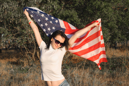 The girl with the American flag outdoors, stars and stripes flag flutters in the wind, happy...