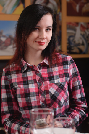 The girl in the plaid shirt looks in a smartphone, a cute brunette with red lipstick on, waiting an order in a coffeeの写真素材