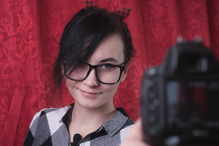 A young female blogger shoots himself on camera, cute brunette, hipser glasses, lifestyle vlog, red backgroundの写真素材