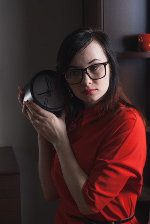 Business woman holding a clock, a portrait of a woman in a red dress, a photo in a dark way, brunette in glassesの写真素材