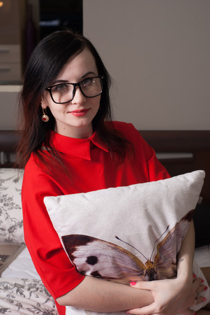 Beautiful girl in red dress and white glasses presses the pillow to his chest, a cute brunette in the bedroomの写真素材