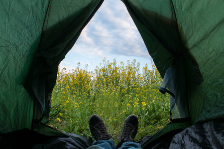 A tourist living in a tent with a view of canola field and a cloudy sky, feet sticking out of the tent, relax in the outdoorの写真素材