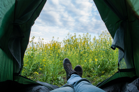 A tourist living in a tent with a view of canola field and a cloudy sky, feet sticking out of the tent, relax in the outdoorの写真素材