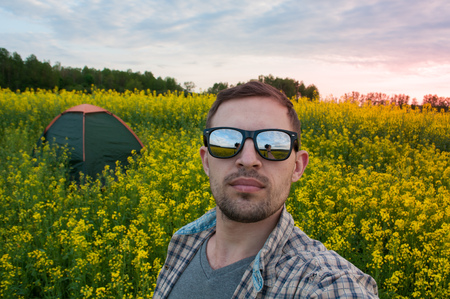 A tourist wearing sunglasses making selfie in the camping,on the background of the tent, rapeseed field at sunsetの写真素材