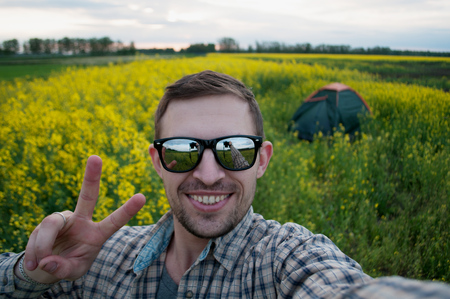 A happy camper in sunglasses making selfie in the camping,on the background of the tent, rapeseed field at sunsetの写真素材