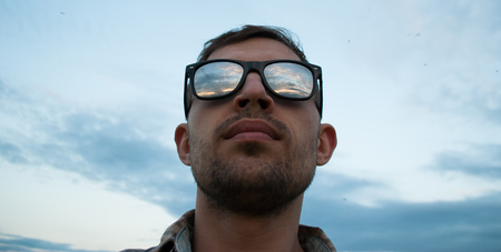 young man in sunglasses on the background of the cloudy sky watching the sunset, the sky is reflected in the glasses of a tourist outdoorの写真素材
