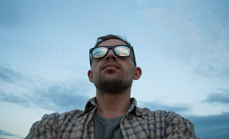 young man in sunglasses on the background of the cloudy sky watching the sunset, the sky is reflected in the glasses of a tourist outdoorの写真素材
