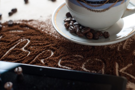 Cup with a hot beverage, roasted coffee beans close-up scattered on the Desk, digital tablet, lettering ground coffee hashtag breakの写真素材