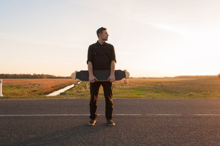 Young man standing on the road with longboard in his hands, guy with longboard at sunsetの写真素材