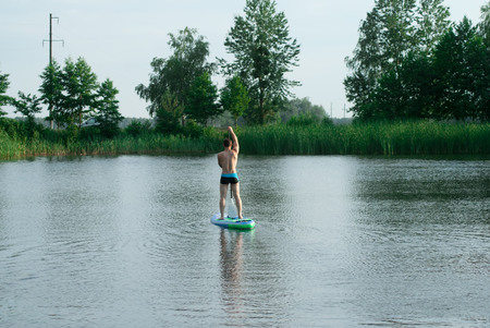 A man stands on a SAP Board with an oar and swims through a forest lake, active rest on the waterの写真素材