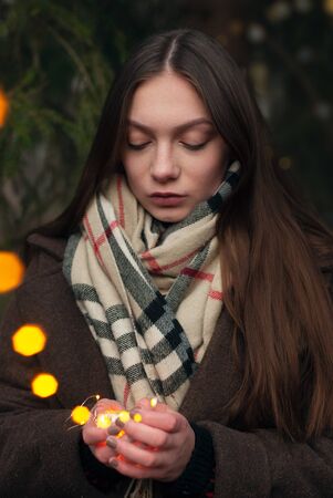 Sad girl in the forest before the new year. Garland with lights. Portrait of a pretty brunette in a frosty Park.の写真素材