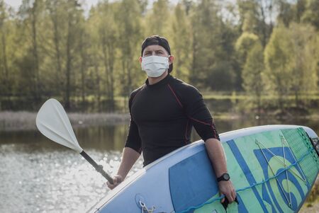 Athletic man in a medical mask stands with a paddle on the background of the lake, paddleboard athlete,の写真素材