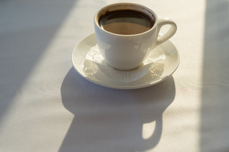 White coffee Cup on a table covered with a tablecloth in a beam of sunlight and long shadowsの写真素材