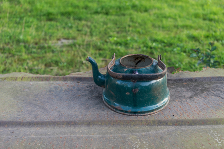 Old enameled kettle on the metal sheet on the background of green grass. For design and networking.の写真素材