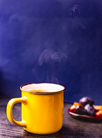 Dark blue background, yellow cup with hot tea on wooden tea stand, and some fruits on a little ceramic plateの写真素材