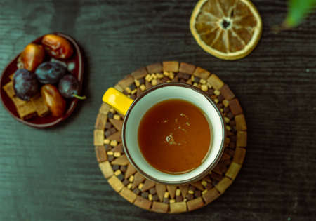 Top view  hot herbal tea in a yellow cup on a black wooden textured table, dried orange slice, brown sugar cubes, and plum fruits, autumn fruits, and healthy teaの写真素材