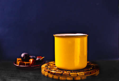 Dark blue background, yellow cup with hot tea on wooden tea stand, and some fruits on a little ceramic plateの写真素材