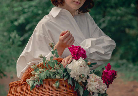 Girl in white clothing with basket of peony flowers in garden. Woman florist hands close up holding summer bouquet. Bohemian or Boho-chic style background with copy space.の写真素材