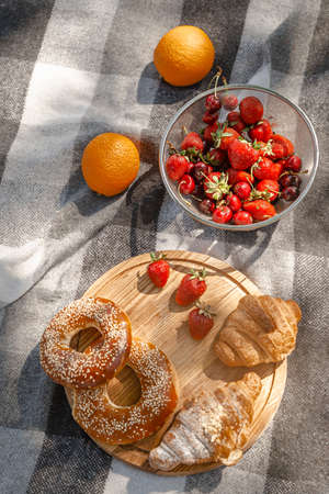 Summer picnic with a basket of Croissants and bagels on rustic wooden desk. French tasty picnic top view, copy space. Strawberries, oranges, cherries in pile. Beach gray black blanket Camping snackの写真素材