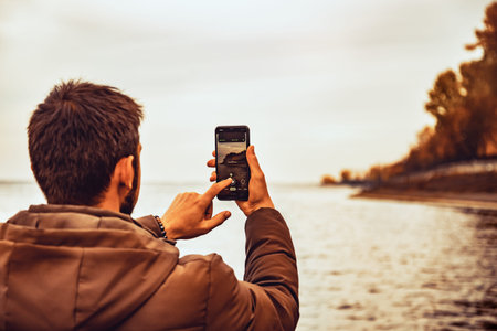 Young male with dark hair in warm jacket taking photo on the shore. Traveler makes mobile...