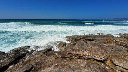 A breathtaking view of vibrant ocean waves crashing against a rugged shoreline under a bright blue...