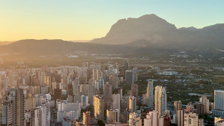 A stunning sunset casts vibrant hues over a bustling city skyline framed by majestic mountains in...