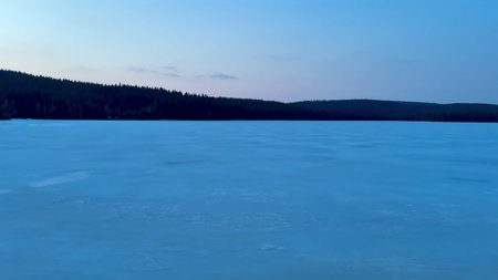 A breathtakingly tranquil view of a completely frozen lake surrounded by tall trees as dusk gently...