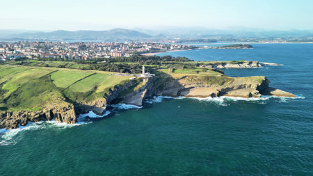 Stunning aerial view of a lighthouse on a rocky coast with greenery and cityscape in the background.の写真素材