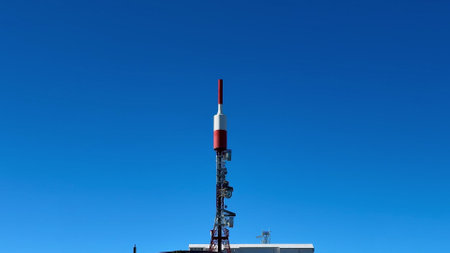 A striking red and white telecom tower prominently stands out on a lush green hillside beneath a bright clear blue skyの写真素材