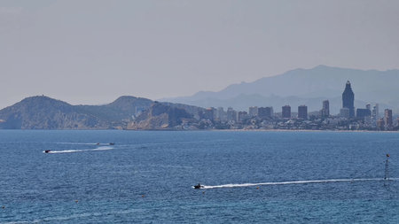 Enjoy a stunning view of a vibrant coastal city with boats gliding over water against majestic mountain backdropsの写真素材