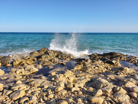 The beautiful ocean waves are crashing powerfully against the rocky coastline beneath a stunning clear blue skyの写真素材