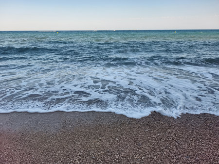 A serene and tranquil view of gentle waves lapping against a smooth pebble beach beneath a clear blue skyの写真素材