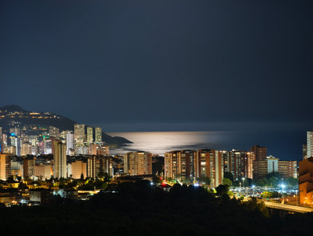 Behold a stunning nighttime view of a coastal city, brightly lit by city lights and ocean reflectionsの写真素材