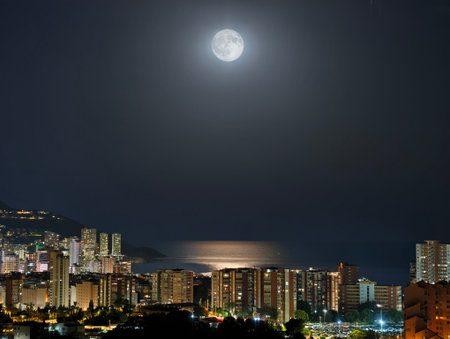 A breathtaking view of a city skyline illuminated by a full moon, beautifully reflecting on the calm sea at nightの写真素材
