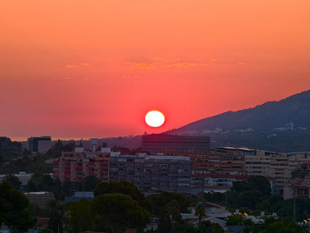A breathtaking sunset is gracefully casting vibrant orange hues across a serene city and adjacent hills at duskの写真素材