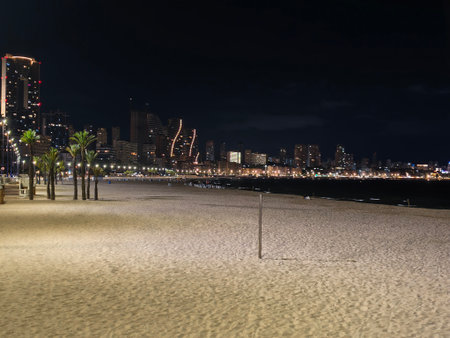 cinematic nocturnal image showcasing illuminated beach with dark waters and skyline in backgroundの写真素材