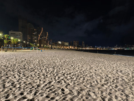 silent beach under city lights, nighttime city beach with rippled sand and distant glowing skylineの写真素材