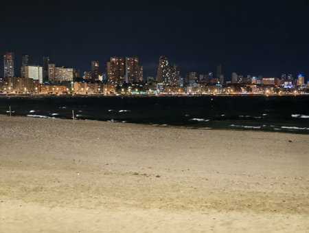 gentle tide lapping dark shoreline with city lights, small breakers, textured sand and calm coastalの写真素材