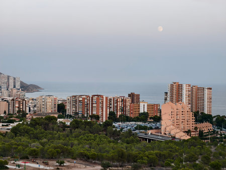 A stunning coastal view showcasing modern buildings and a serene ocean scene at dusk complemented by the moonの写真素材