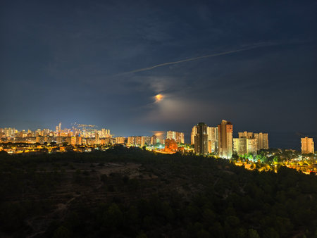 The stunning nighttime view of an urban skyline is lit up by vibrant city lights under a glowing moonの写真素材