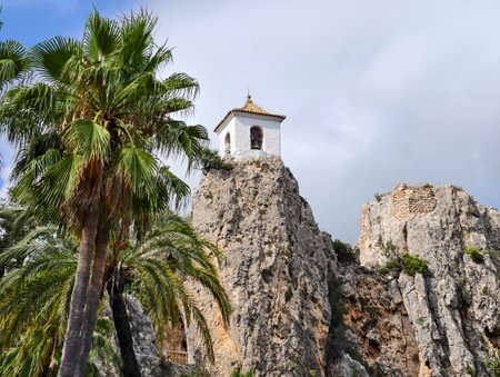 tropical bell tower, scenic scene of white tower on rocky terrain surrounded by palms and cloudsの写真素材