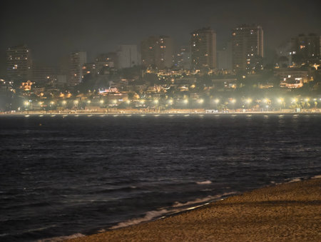 coastal night with city illumination, dark ocean waves crash on sandy shore as city glows nearbyの写真素材