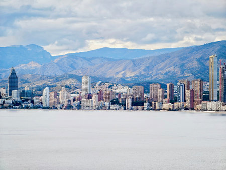 city skyline framed by distant mountains, soft daylight filtering through clouds, calm bay edge, modern towersの写真素材