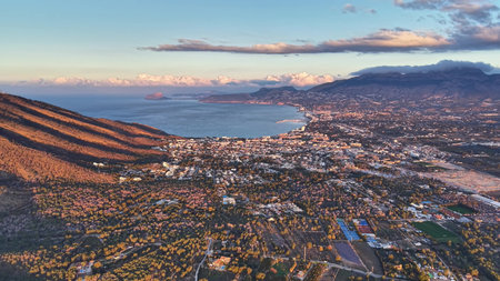 Sunset aerial revealing bay rolling ridgeline sprawling town, warm highlights on slopes and reflective waterの写真素材
