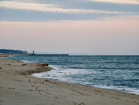 Sunset coastline with distant lighthouse silhouette and gentle surf, pastel sky and empty sand offer