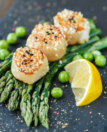 Scallops with sesame seeds, asparagus, lemon and green peas on a black plate. Selective focusの写真素材